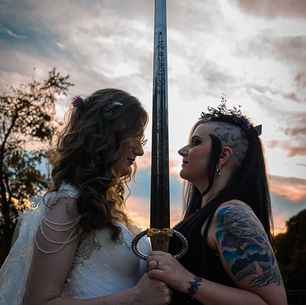 A fierce and romantic couple, one in a white gown and the other in a black dress with a crown, stand face to face holding a sword between them at sunset.