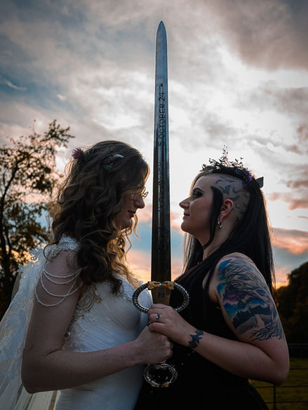 Ethereal gothic wedding portrait of two brides holding a sword under a dramatic sky, blending alternative and unique photography style.