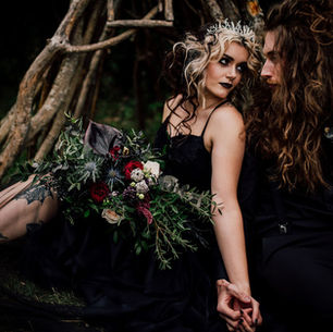A darkly romantic couple in gothic wedding attire sit hand in hand, surrounded by nature, with the bride holding a moody bouquet of deep red and black florals.