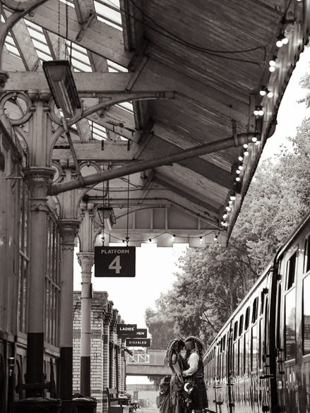 Whimsical alternative wedding couple sharing a romantic moment at a vintage train station under an aged platform canopy.