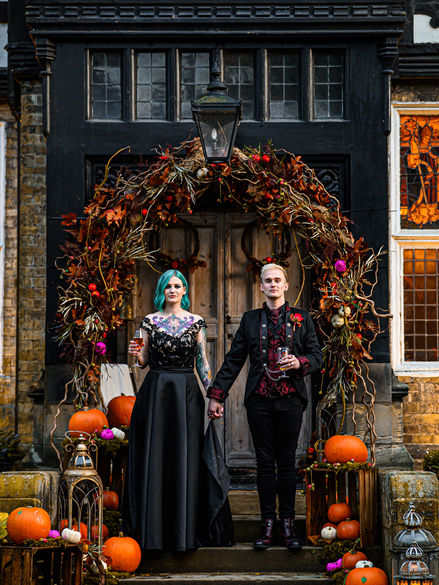 Gothic wedding couple stands in front of an autumnal arch adorned with pumpkins and foliage, showcasing alternative style.