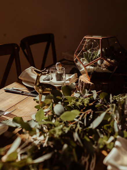 Alternative wedding table with rustic styling, featuring greenery, a glass terrarium centerpiece, wooden details, and soft lighting.
