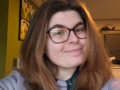 Zoe is wearing a gray sweater over a green plaid shirt with a pendant necklace, seated indoors with green and beige walls and framed artwork in the background.