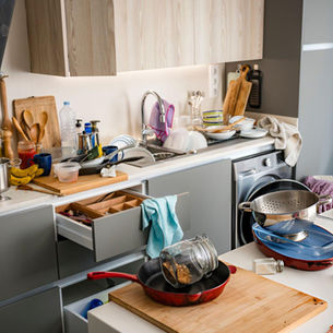A cluttered kitchen bench with dishes, cookware, and open drawers during daily use.