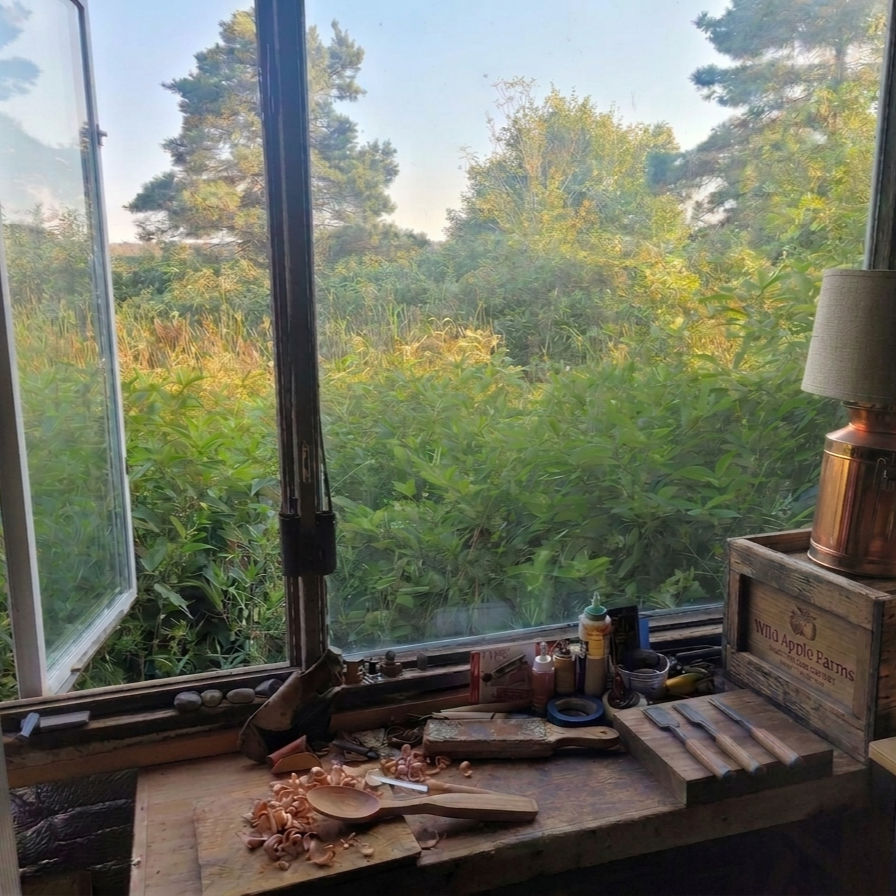 Harald's sunlit woodworking workshop with hand-carved spoons on the workbench, a large window overlooking green forest scenery, and a copper lamp on a wooden box labelled Wild Apple Farms to the right.