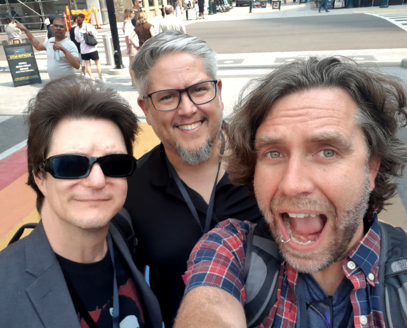 Three men take a street selfie downtown; one shouts playfully, two smile, with pedestrians and city buildings behind them.