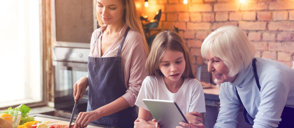 Drei Generationen – Großmutter, Mutter und Tochter – stehen in einer Küche und bereiten gemeinsam Gemüse zu. Die ältere Frau zeigt dem Kind ein Tablet. Symbolbild für digitale Helfer im Haushalt, bewussten Konsum und gemeinsames Sparen mit KI.