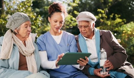 Eine Pflegekraft zeigt zwei älteren Frauen auf einem Tablet neue Möglichkeiten der digitalen Kommunikation – Symbolbild für Leben mit Sinn und KI.