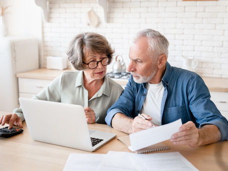 Ein älteres Paar sitzt gemeinsam am Tisch. Die Frau tippt auf einem Taschenrechner, der Mann hält ein Dokument und schaut konzentriert auf den Laptop. Symbolbild für Finanzschutz, digitale Sicherheit und den Einsatz von KI im Geldmanagement.