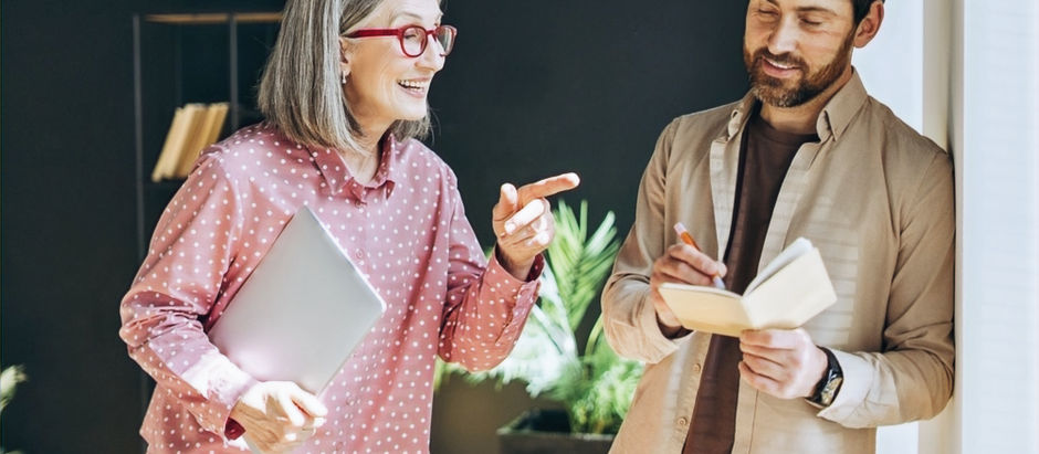 Eine ältere Frau mit Tablet spricht lächelnd mit einem jüngeren Mann, der in ein Notizbuch schreibt. Beide wirken konzentriert und freundlich. Symbolbild für Austausch, Verständnis und digitale Demokratie mit KI-Unterstützung.