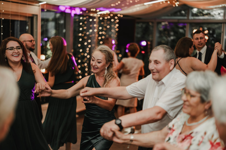 Fotograf ślubny Poznań- Uśmiechnięci goście tańczący w kółku na przyjęciu weselnym z lampkami.