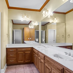  A spacious bathroom featuring a large L-shaped vanity with a white countertop, dual sinks, and wooden cabinets. The vanity is complemented by an expansive mirror with multiple light fixtures mounted above. The walls are painted in a warm beige color, with wooden crown molding adding contrast. To the left, there is a glass-enclosed shower with a frosted door. The floor is covered in pinkish-beige tiles, and a doorway in the background leads to another section of the home.