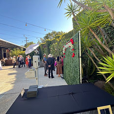Capture the charm of your event with BeütifulPixels' state-of-the-art photo booth, shown here at Stacy and Greg's wedding reception at The Smog Shop in Los Angeles. The booth, prominently placed against a decorative green wall with tropical and floral accents, offers guests a unique and engaging entertainment option. Complete with a professional lighting setup and a nearby table displaying beautifully crafted photo print keepsakes, our booth ensures every moment is captured in perfect clarity. This image highlights the inviting and interactive experience provided by our photo booth, making it an essential addition to any celebration.