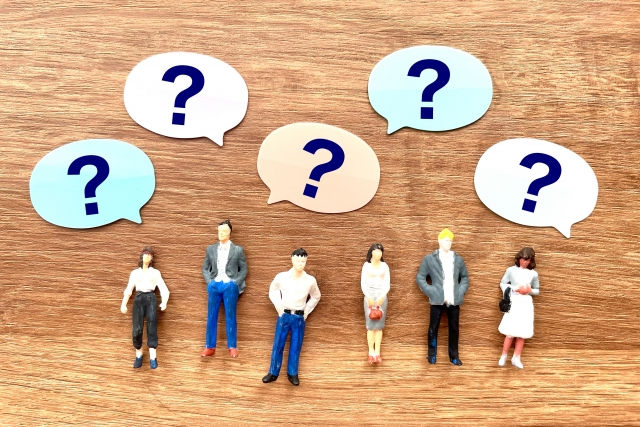 木目の背景に、複数のミニチュア人物と色違いの吹き出しに?が並び、相談や疑問を示すような場面