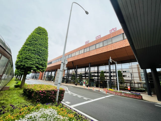 赤いレンガ調の駅舎前に、剪定された木や花壇、街灯が並ぶ静かな道路。屋上に秋田空港の文字が見える。