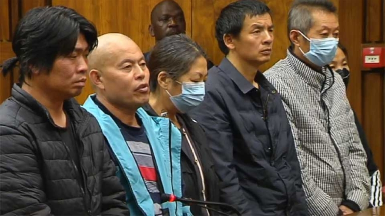 Five people stand in a courtroom setting, some wearing masks. They appear serious and attentive. Wood paneling is visible in the background.
