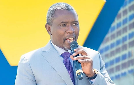 Kudakwashe Tagwirei. Man in a gray suit speaks into a microphone against a bright blue and yellow geometric background. He appears focused and engaged.
