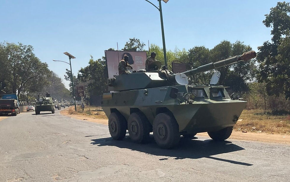 Military vehicles on a road with soldiers on top, surrounded by trees and a billboard. Clear sky and a calm atmosphere.
