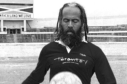 Man with braided hair, wearing a "Toronto!" shirt, balances a soccer ball. Background shows a stadium with "Jamaica Land We Love" text.