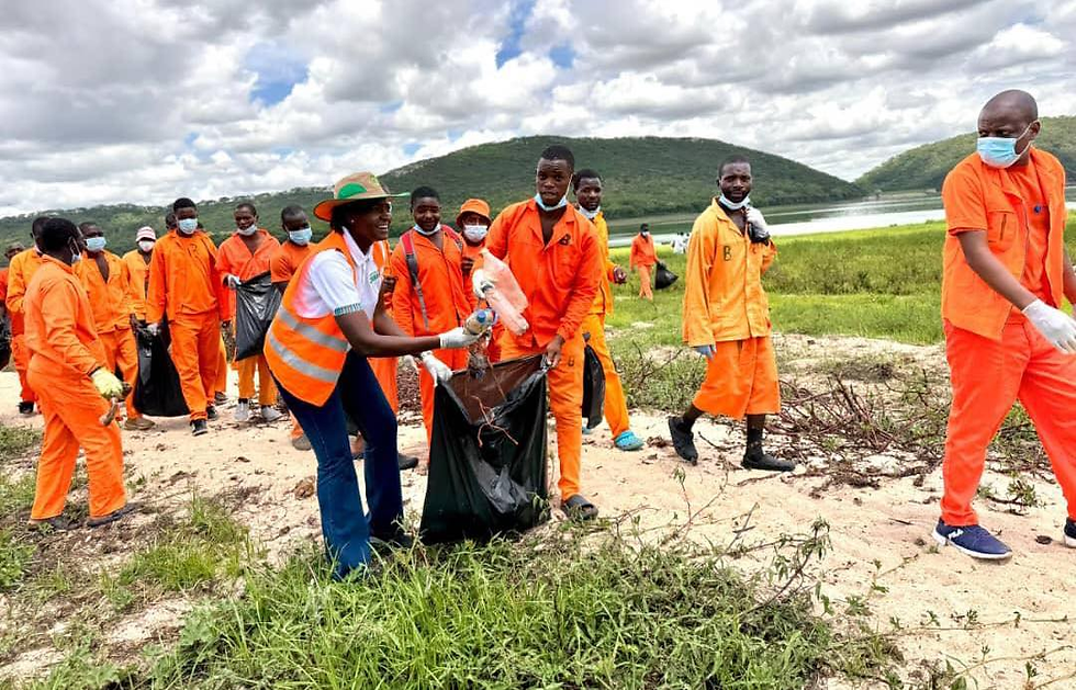 People in orange uniforms and masks clean up trash near a lake, with green hills in the background under a cloudy sky.