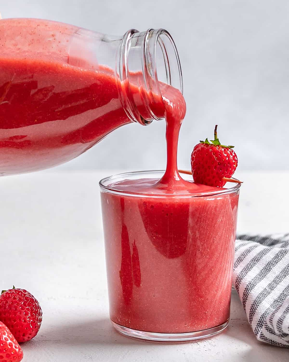 A refreshing summer smoothie being poured alongside a scattered bowl of nutrient-dense fresh strawberries.