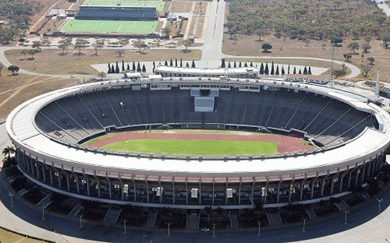 Aerial view of an empty oval stadium with green field and running track. Surrounding are roads and fields, with trees scattered around.