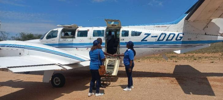 Three people in blue shirts stand by a small white plane with blue stripes, on a dirt runway, under a clear blue sky, engaging in conversation.