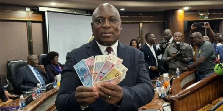 Man in suit displays colorful banknotes at a conference. People in business attire and photographers surround him. Room has wood paneling.