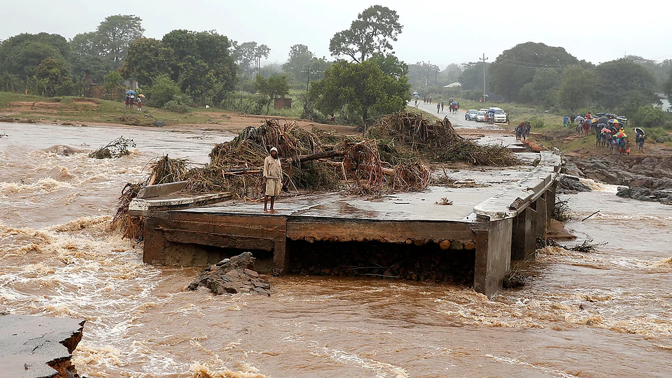 Person stands on a damaged bridge amid floodwaters. Debris piles up; people with umbrellas walk nearby. Overcast sky sets a somber mood.