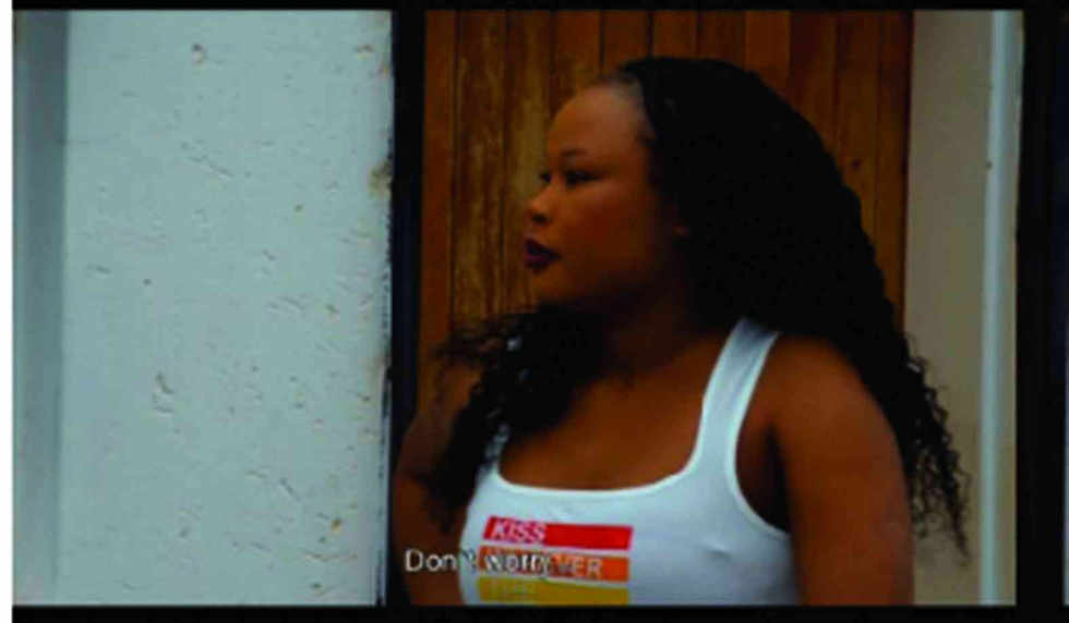 Woman in white tank top with colorful text, standing against a wooden door and white wall, looking to the side. Subtle mood.