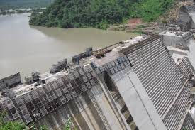 Concrete dam construction site overlooking a river, with lush green hills in the background, under a clear sky. No visible text.