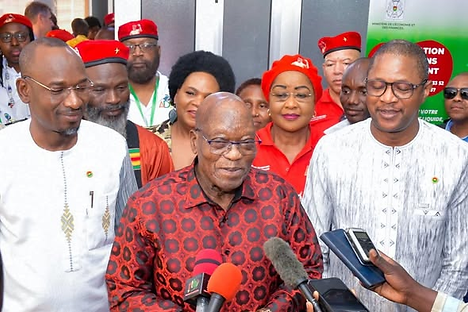 Group of people in colorful attire, smiling at a press event. Microphones are visible in the foreground. The mood is cheerful and vibrant.