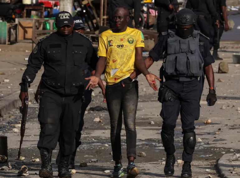 Police escort a man in a yellow shirt with raised hands through a debris-strewn street. Tension is evident. Officers wear dark uniforms.