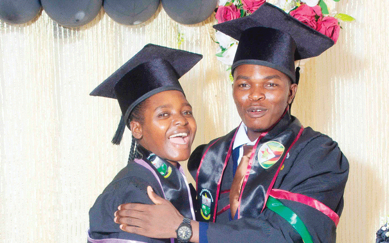 Two graduates in black gowns and caps embrace joyfully. Background has balloons and flowers. They're smiling, conveying a celebratory mood.
