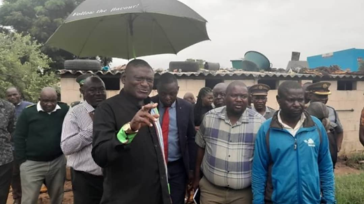 Group of men outdoors; one holding an umbrella with "Follow the flavour!" text. Overcast sky, corrugated building roof behind them.