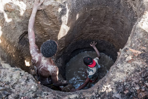 Two people digging a deep muddy hole, one covered in mud, the other reaching out. Brown dirt walls and water at the bottom, outdoors.