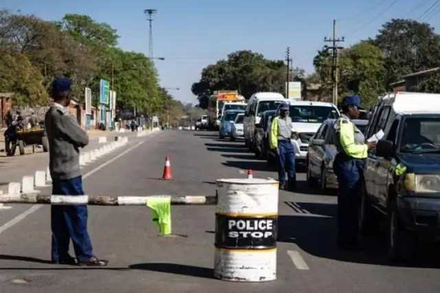 Police checkpoint on a road with officers inspecting cars. "Police Stop" on a barrel. Trees and vehicles line the street.