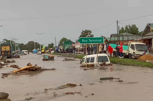 Flooded street with partially submerged vehicles, debris, and people in the distance. A green "Bus Terminal" sign is visible. Overcast sky.