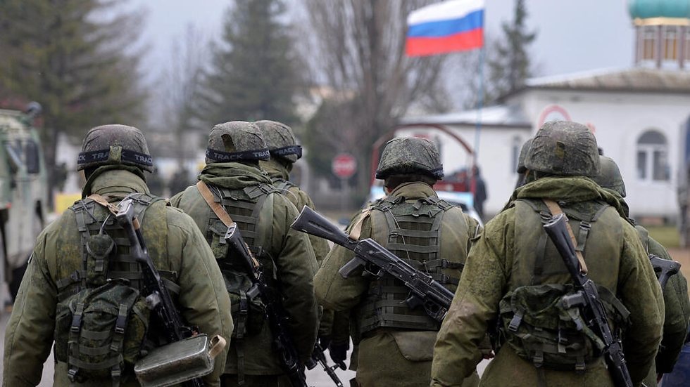 Soldiers in green camouflage and helmets walk with rifles in a street. A Russian flag waves in the background on a cloudy day.