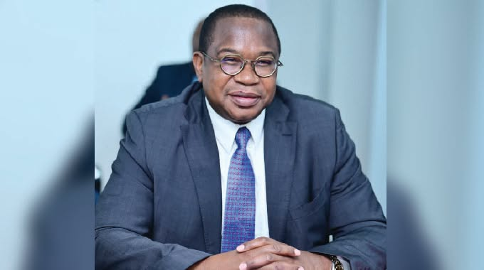 Man in a suit and tie sits at a desk, smiling. He wears glasses and is in a bright, professional setting. Neutral background.