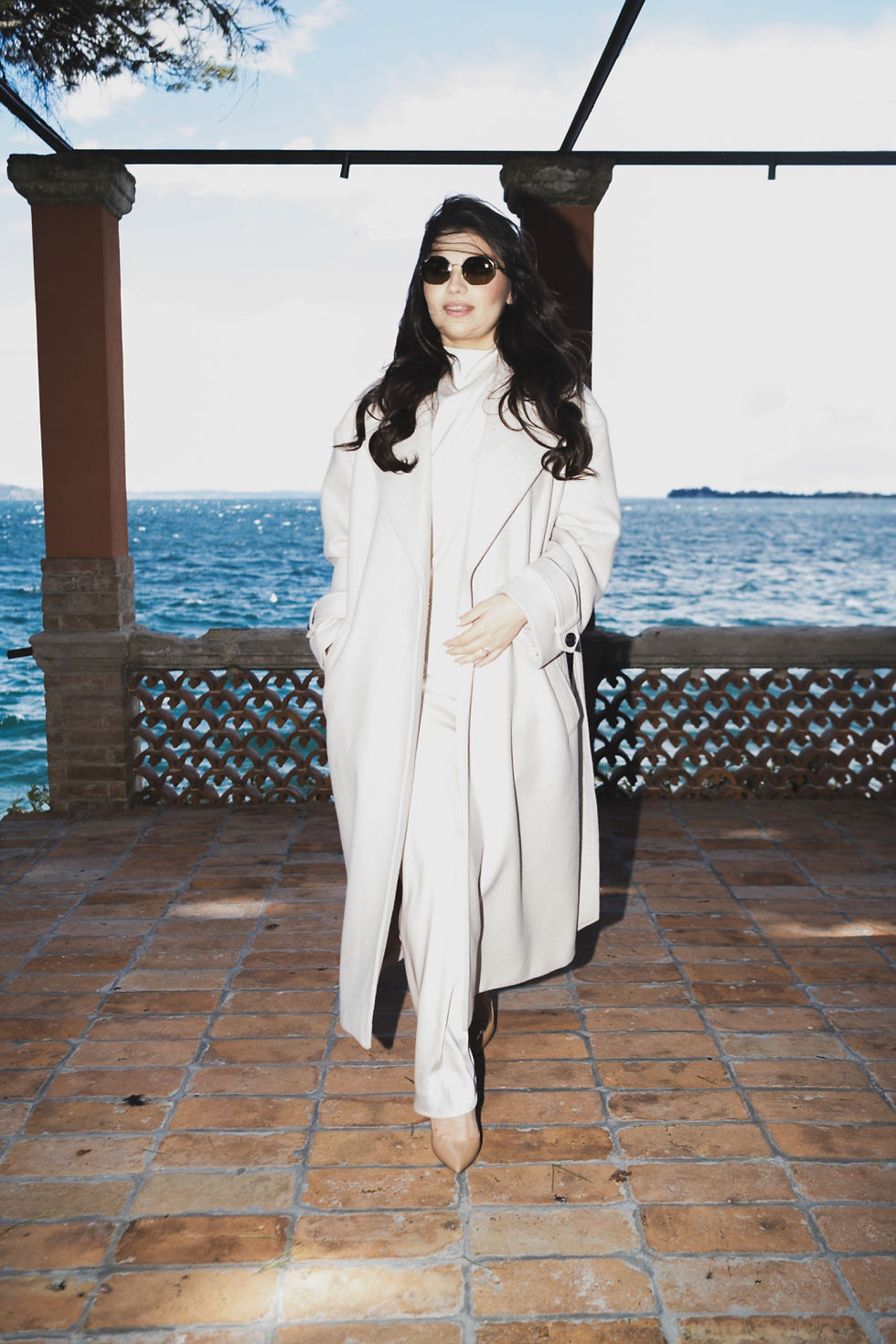 In the luminous flash-kissed light of Lake Garda, a woman in a flowing white coat and elegant sunglasses stands poised on a seaside terrace, her hair gently windblown by the Mediterranean breeze. Behind her, the vivid azure waters merge seamlessly with the clear sky, creating a timeless cinematic backdrop of pure Italian elegance. Captured during the Dolce Vita-inspired editorial photoshoot with AC Fotografia, this refined moment blends effortless style, authentic presence, and classic La Dolce Vita charm. With the iconic vintage red Fiat 500 waiting nearby, every detail from the natural wind-swept movement to the precise balance of ambient light and flash contributes to a sophisticated visual story of freedom, romance, and enduring Italian heritage.