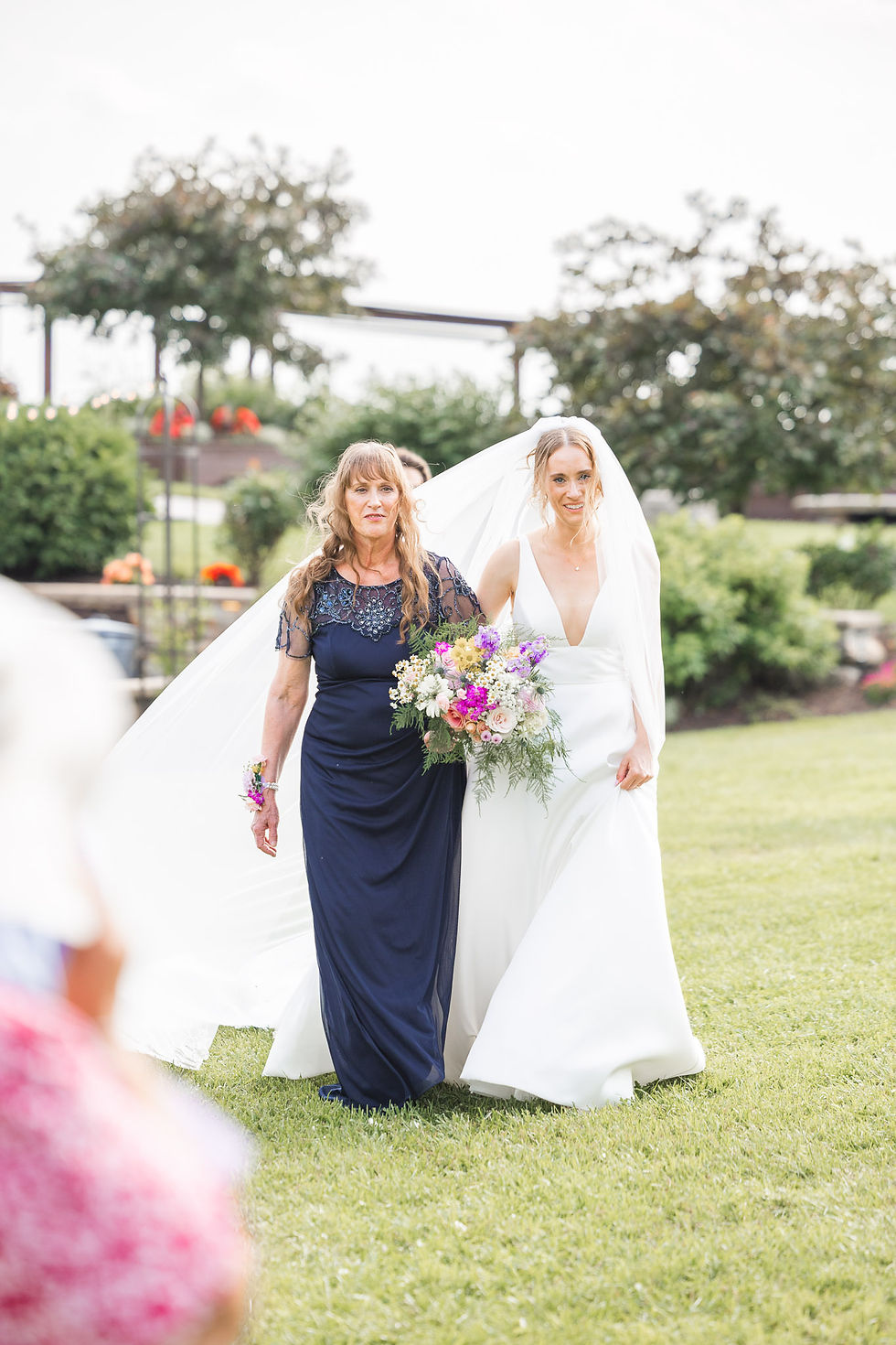 Bride in white, holding bouquet, walks with woman in navy dress on green lawn. Trees and flowers in background. Joyful mood.
