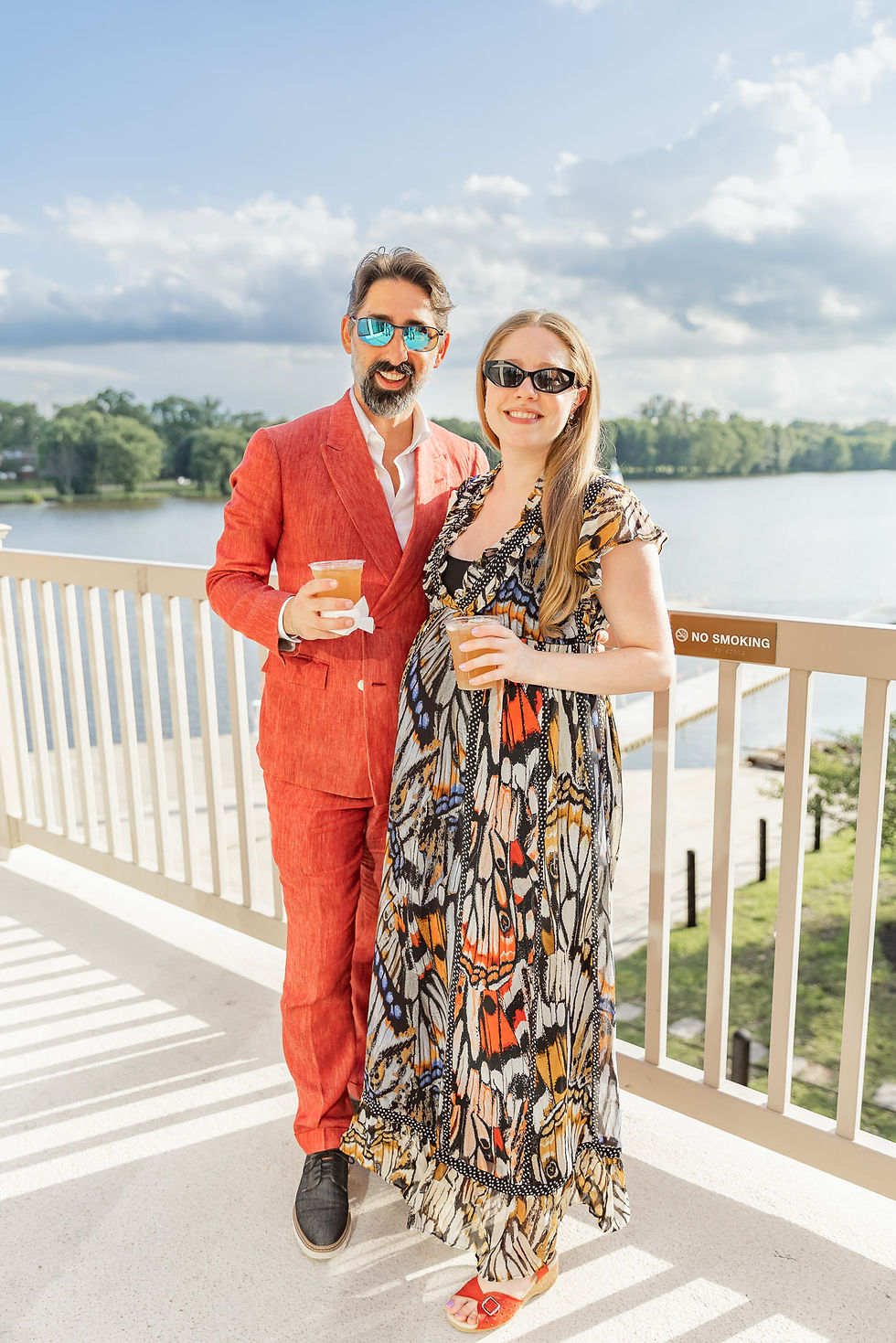 Smiling couple in sunglasses holding drinks on a sunny lakeside balcony; man in coral suit, woman in butterfly-print dress, no smoking sign