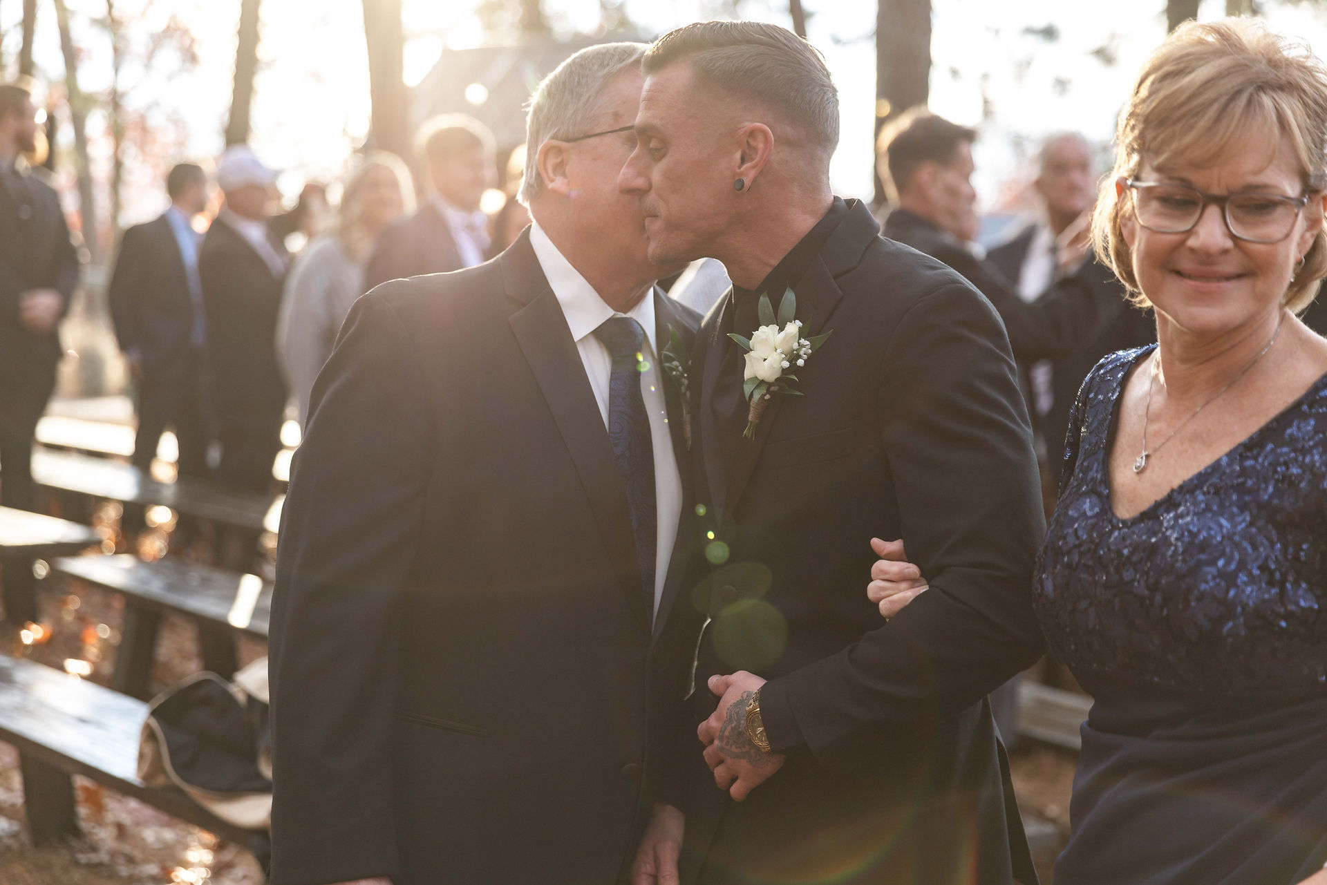 Man in black suit kissses man on the cheek while woman in blue dress holds his elbow with sunflare on the lens from sunset