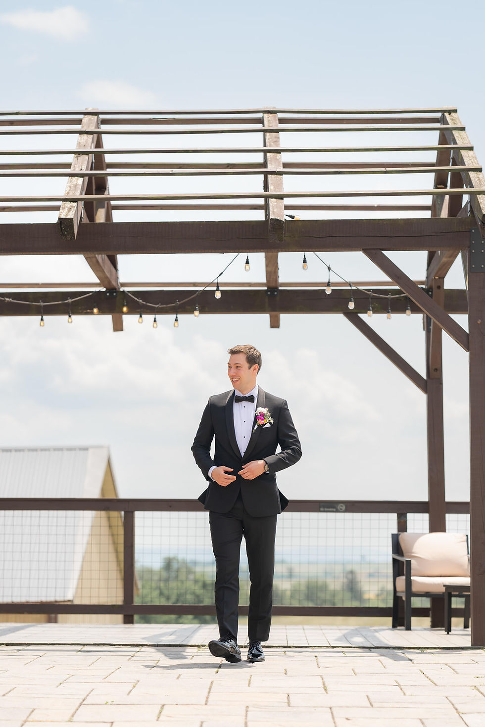 Man in a black suit with a boutonniere walks under a wooden pergola on a sunny day. Bright, open sky and a scenic view in the background.