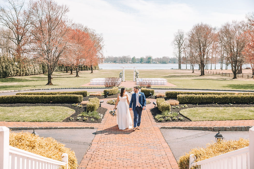 A couple walks hand in hand on a brick path in a garden. Trees with red leaves and a lake are in the background, creating a serene scene.