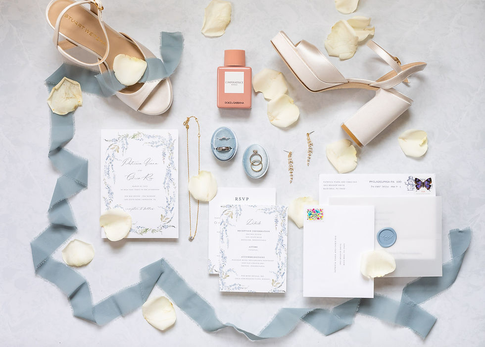 Elegant wedding flatlay with white heels, perfume, blue ribbons, rings, invites, and petals on white background. Inviting, romantic vibe.