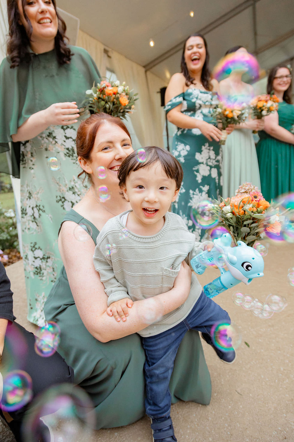 Smiling woman holds a young boy amid bubble-filled celebration, with bridesmaids in green floral dresses and bouquets behind them.