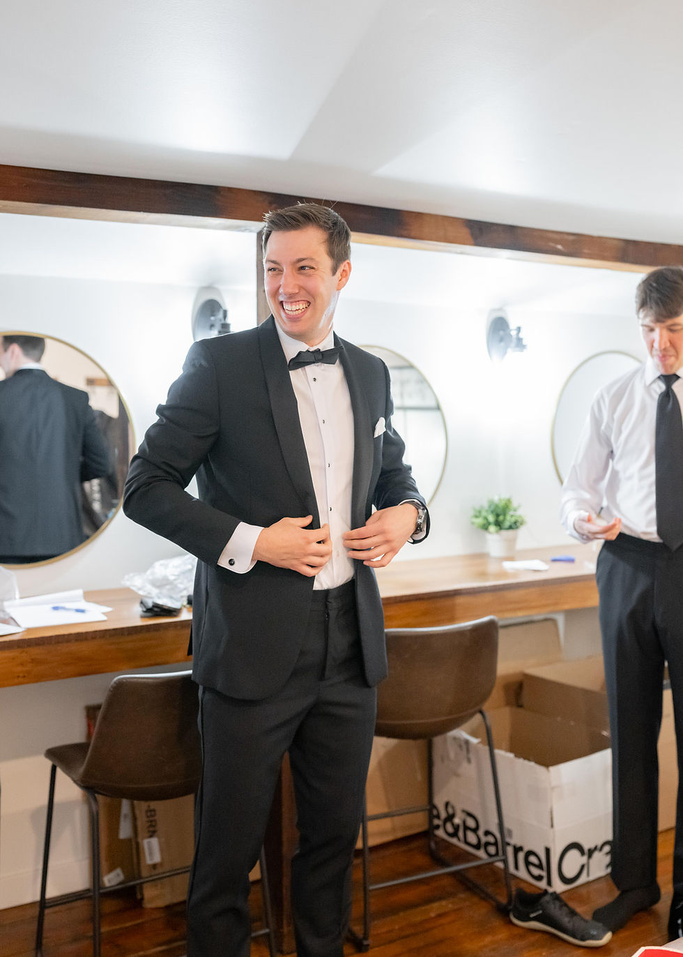 Man in black tuxedo smiling, adjusting jacket, in dressing room with mirrors and wooden counter. Another man fixes tie, box on floor.