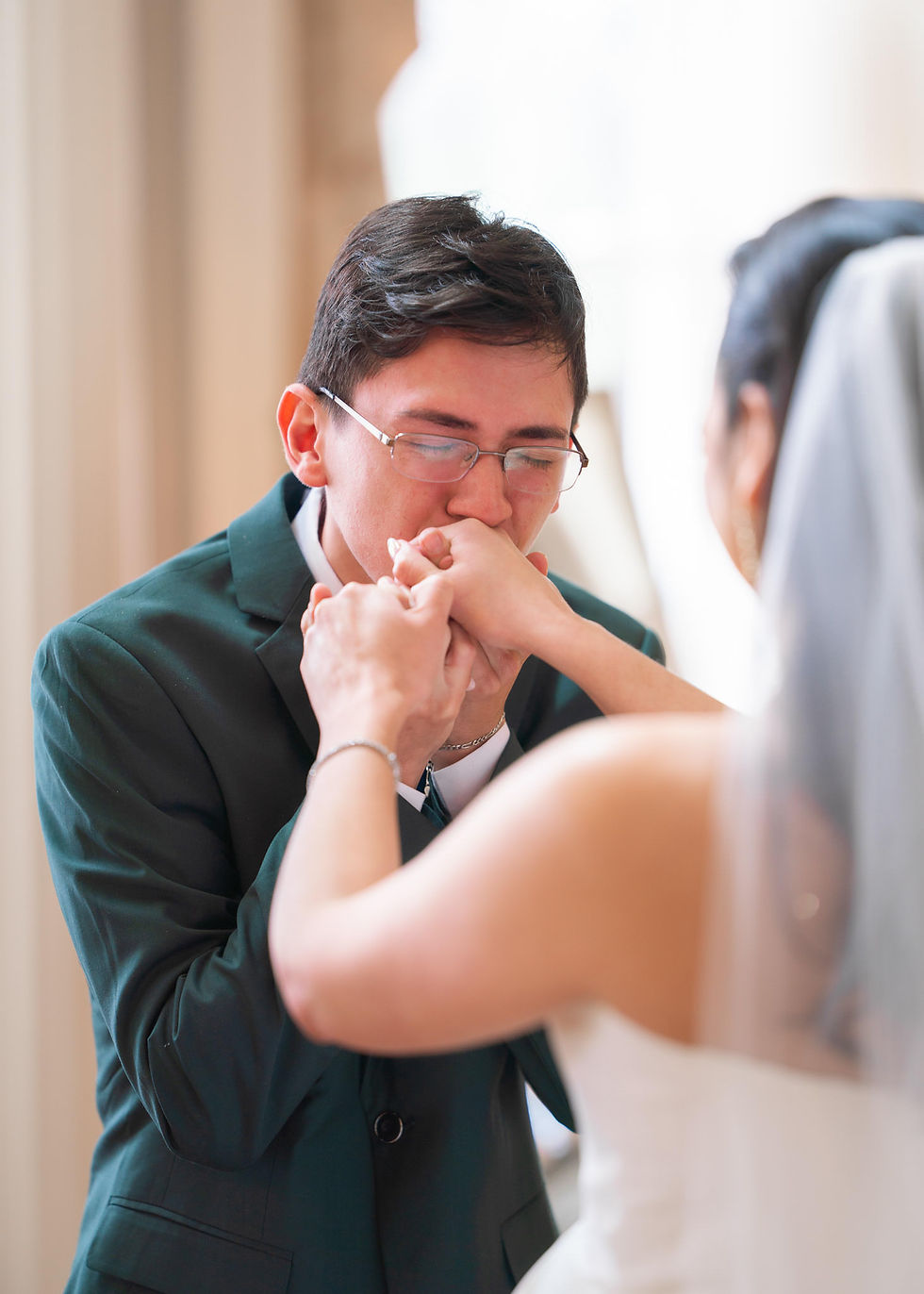 Groom in glasses kisses bride’s hand tenderly, both in wedding attire. Soft lighting, indoor setting, emotional and intimate moment.